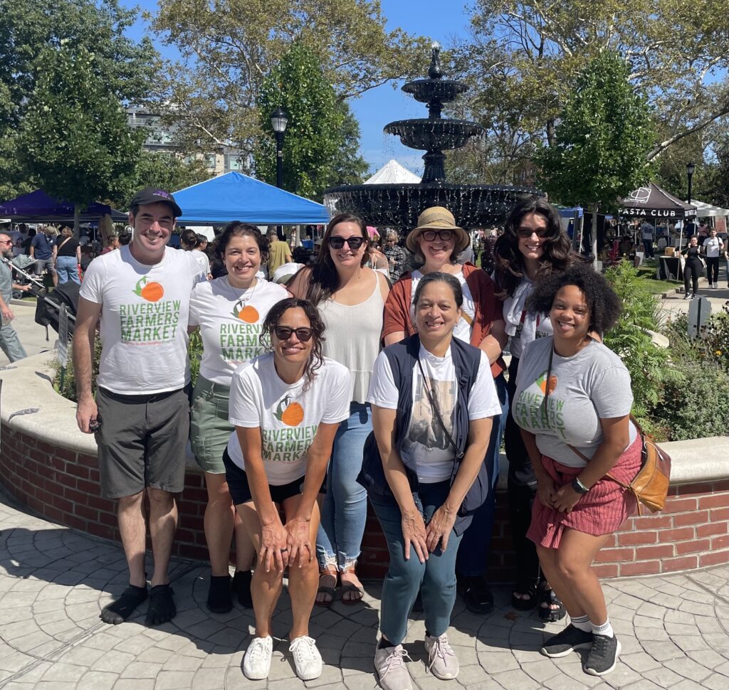 The all-volunteer board of Farms in the Heights stands in front of the Riverview Park fountain on a sunny market day.