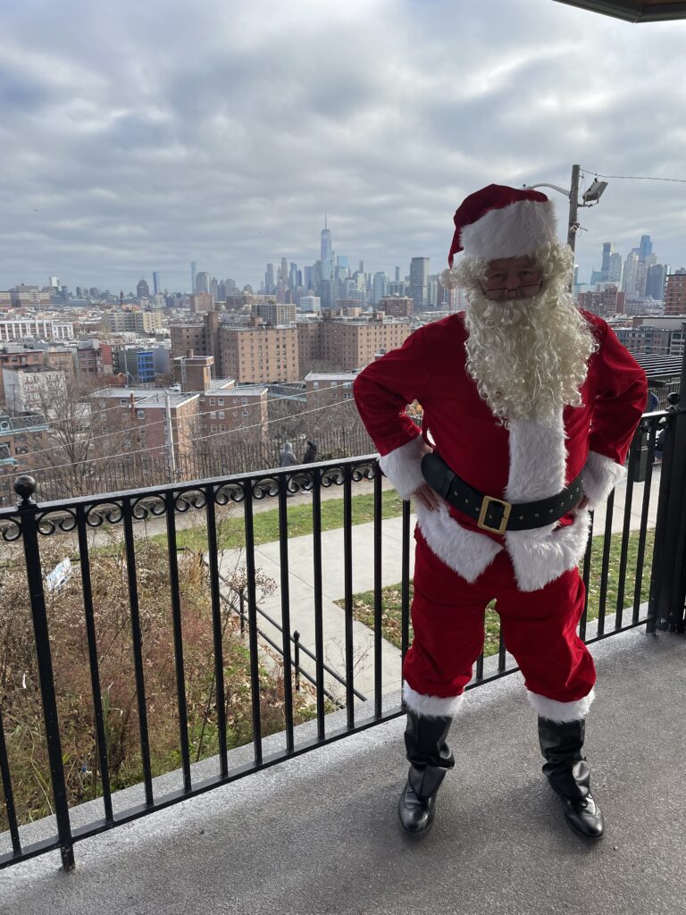 Santa Claus stands with his hands on his hips in front of the New York City skyline from the gazebo in Riverview Park at the 2022 Heights Holiday Market.