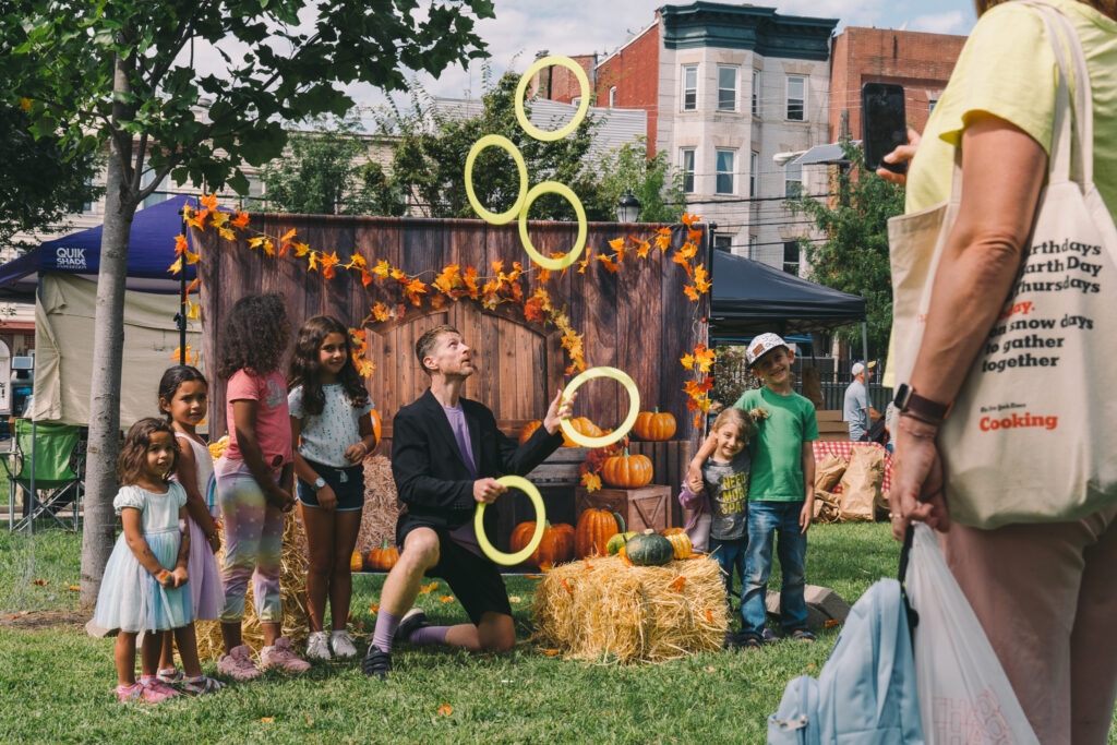 Juggler Sean Blue juggles five neon yellow hoops in front of a fall-themed photo backdrop surrounded by children.