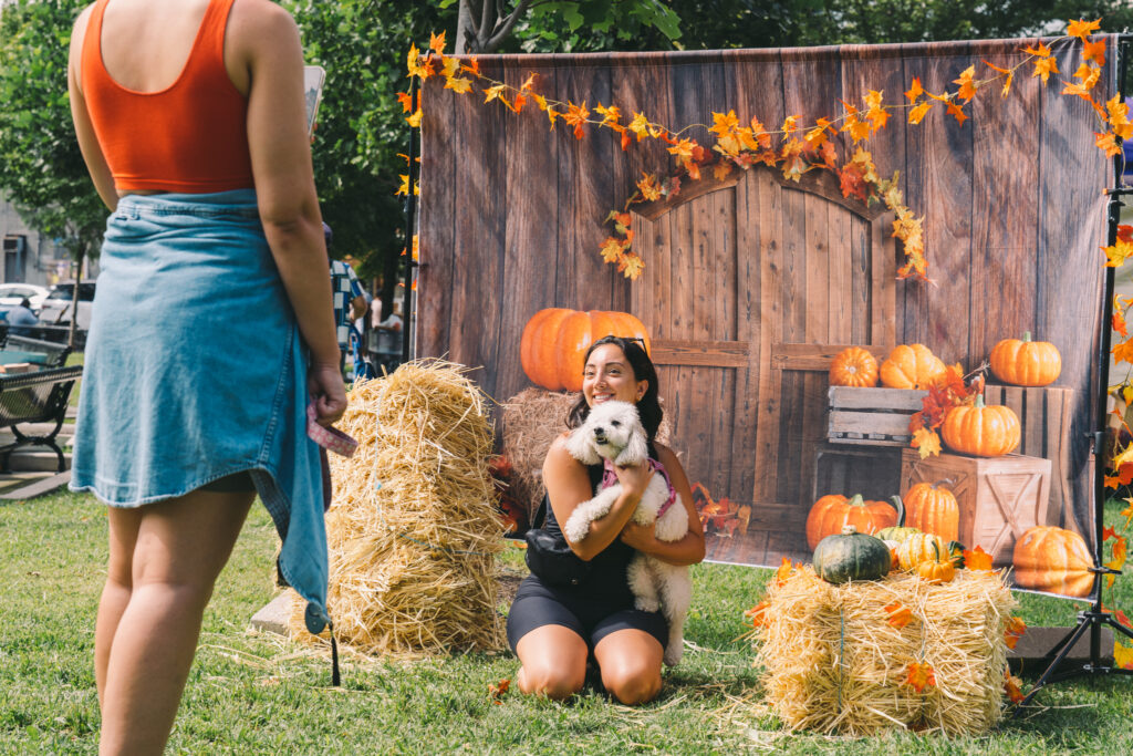 A young woman poses in front of a fall-themed photo backdrop with her fluffy white dog.