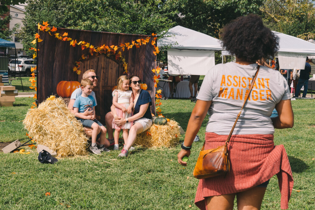 A family of four sits on the hay bales in front of the fall photo backdrop while another person takes their photo.