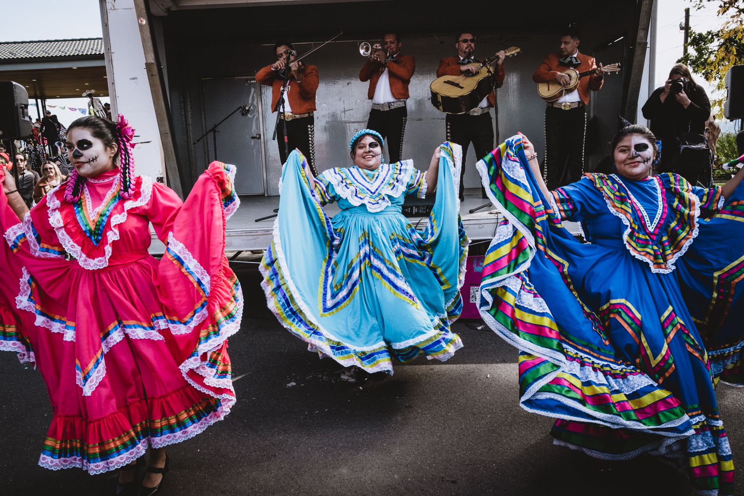 Three dancers dressed in traditional Mexican folk dresses display their colorful skirts in front of a mariachi band at the Riverview Farmers Market's Day of the Dead celebration.