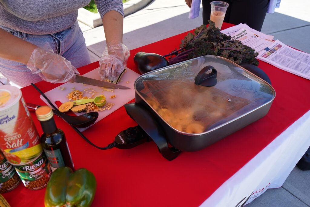 A woman's hands are scene chopping squash on a cutting board with an electric skillet full of steam cooking up something delicious in front of them. She's running a cooking demo at the Riverview Farmers Market.