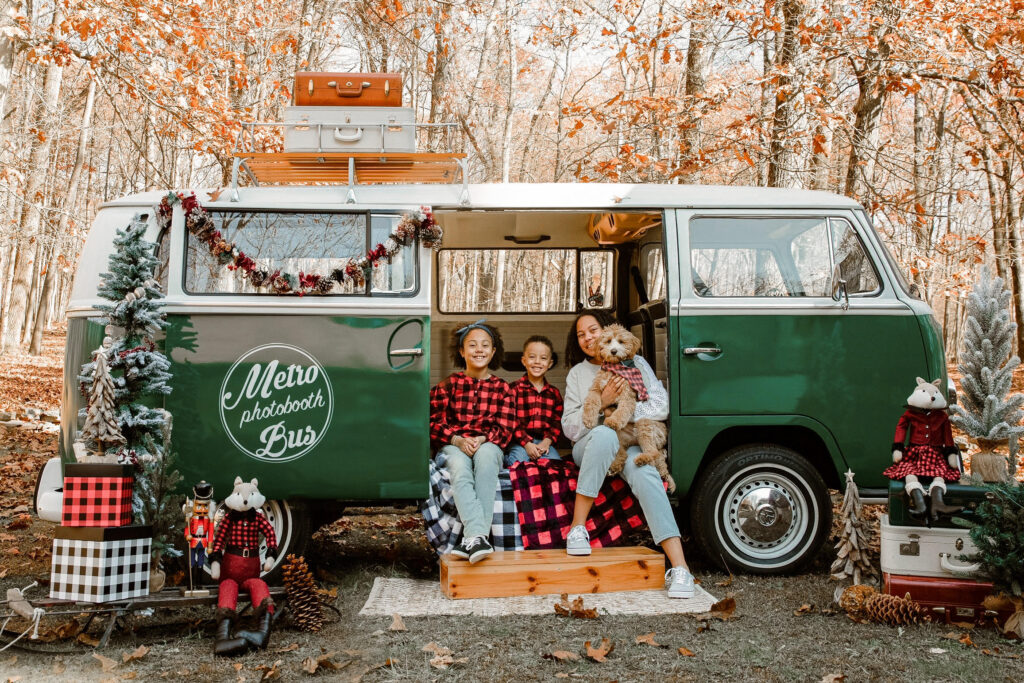 A family in red plaid pajamas sits in the open door of a dark green vintage VW bus with their dog. The sides of the bus are decorated with holiday flair, clearly staged for the photo. On the VW bus is a logo of Metro Photobooth Bus.