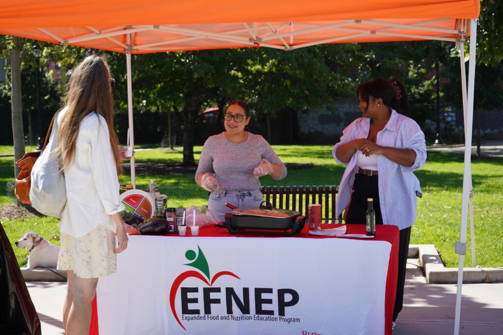 Photo of two women doing a food cooking demonstration at the Riverview Farmers Market. They are from the EFNEP Program at Rutgers. They are speaking to a third person.