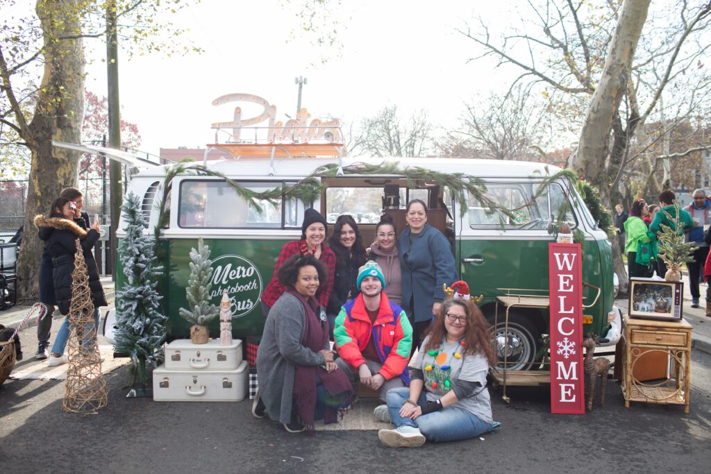 Photo of a group of Farmers Market board members and staff in front of a Volkswagen bus that has been converted into a photo booth - at the Holiday Market 2023