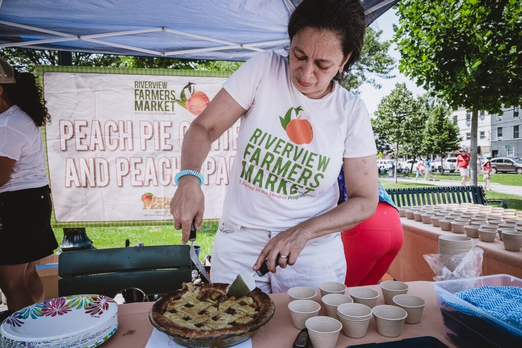 Photo of a woman cutting up a pie for the judges to taste at the 2023 Peach Pie Contest at Riverview Farmers Market. There is a banner behind her.