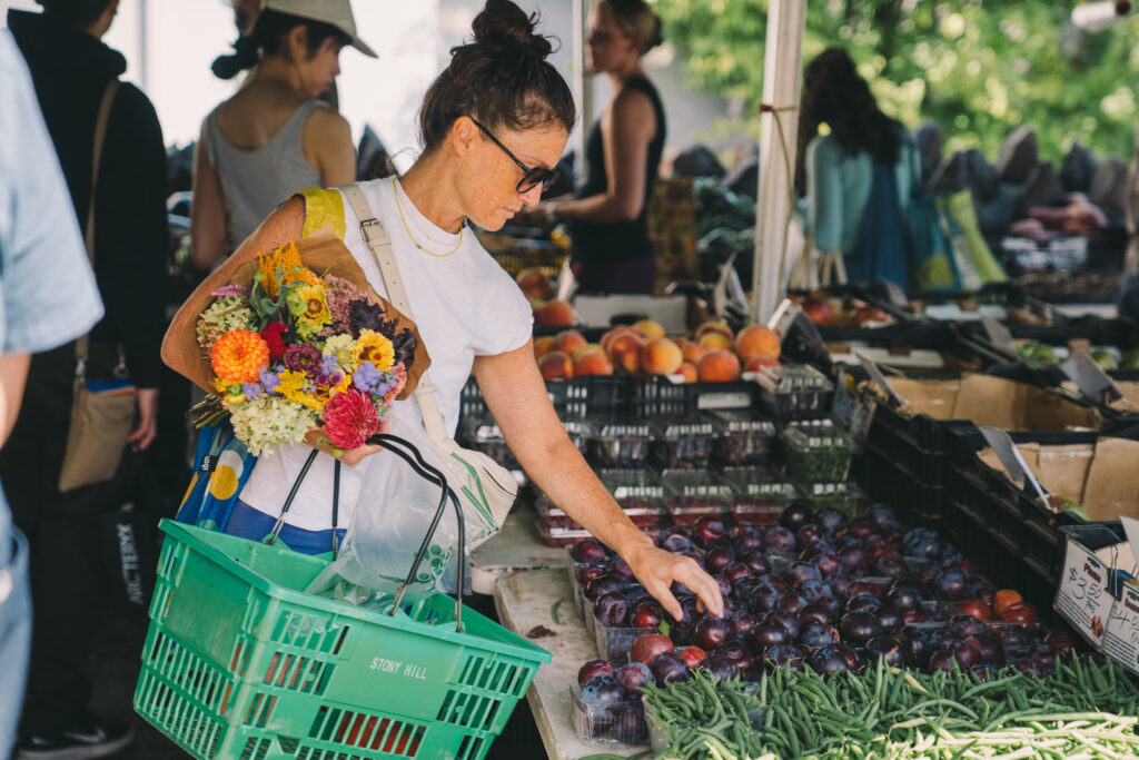 Photo of a white woman shopping at a farmers market tent. She is wearing a white t-shirt, jeans and sunglasses. Her hair is up in a bun. She is holiding flowers, and picking out plums from a table. 