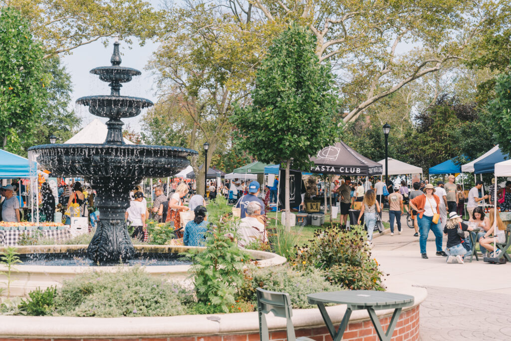 Photo of multiple farmers market vendor tents surrounding a running fountain on a summer day. Green trees and park can be seen in the background