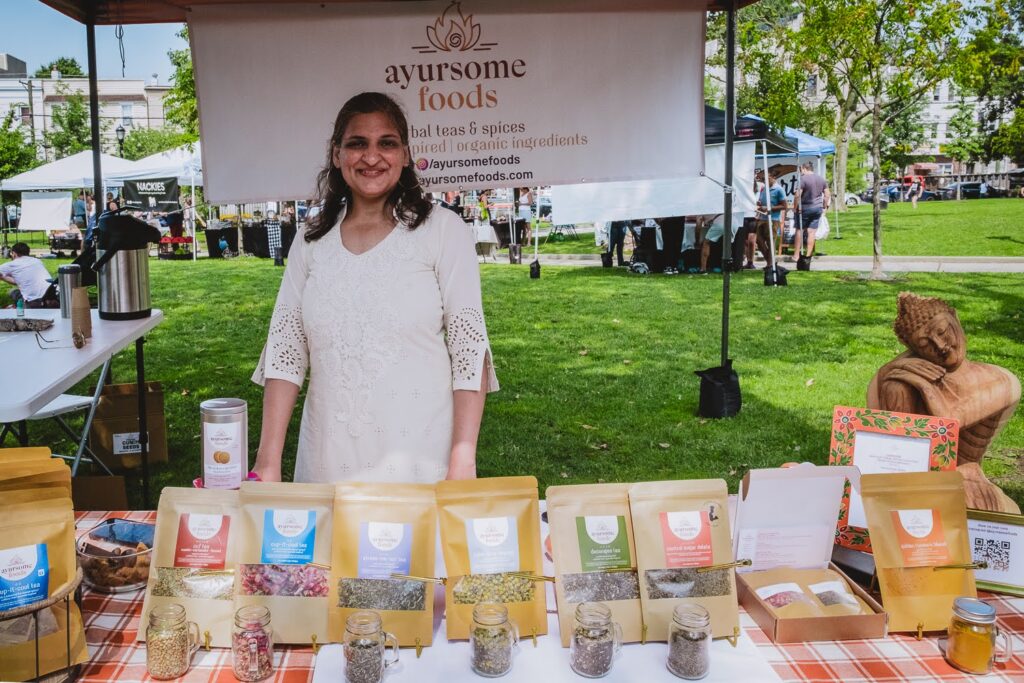 Photo of a smiling South Asian woman, wearing a white tunic top, in a farmers market booth on a summer day, selling tea.