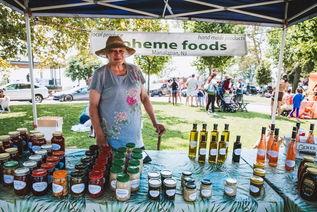 Photo of a smiling White woman, wearing a floral t-shirt and a large sun hat, in a farmers market booth on a summer day, selling pickled and fermented items.