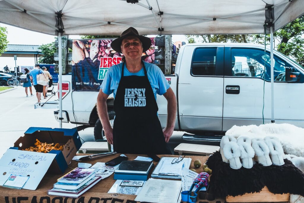 Photo of an White man, wearing a blue t-shirt and black apron, in a farmers market booth on a summer day, selling lamb and lambskin products