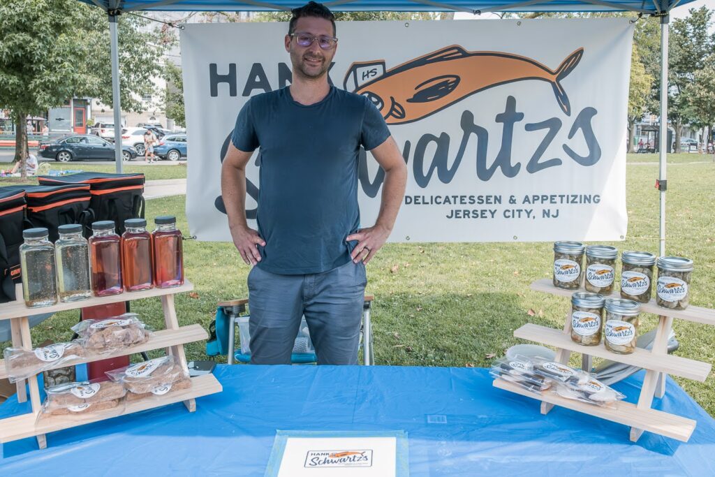 Photo of an White man in a blue t-shirt and jeans, in a farmers market booth on a summer day, selling Jewish food