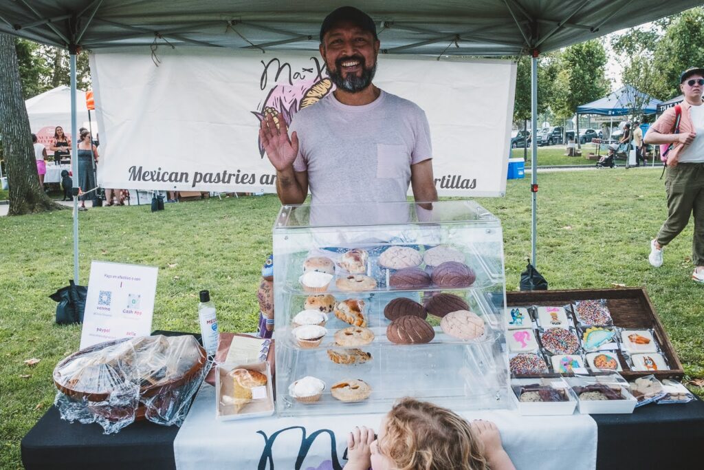 Photo of a smiling Hispanic man, in a white tee shirt, in a farmers market booth on a summer day, selling Mexican pastries