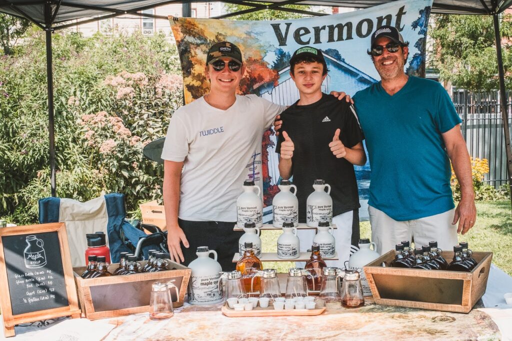 Photo of a two White men and a White teen, smiling at the camera, in a farmers market booth on a summer day, selling Vermont maple syrup