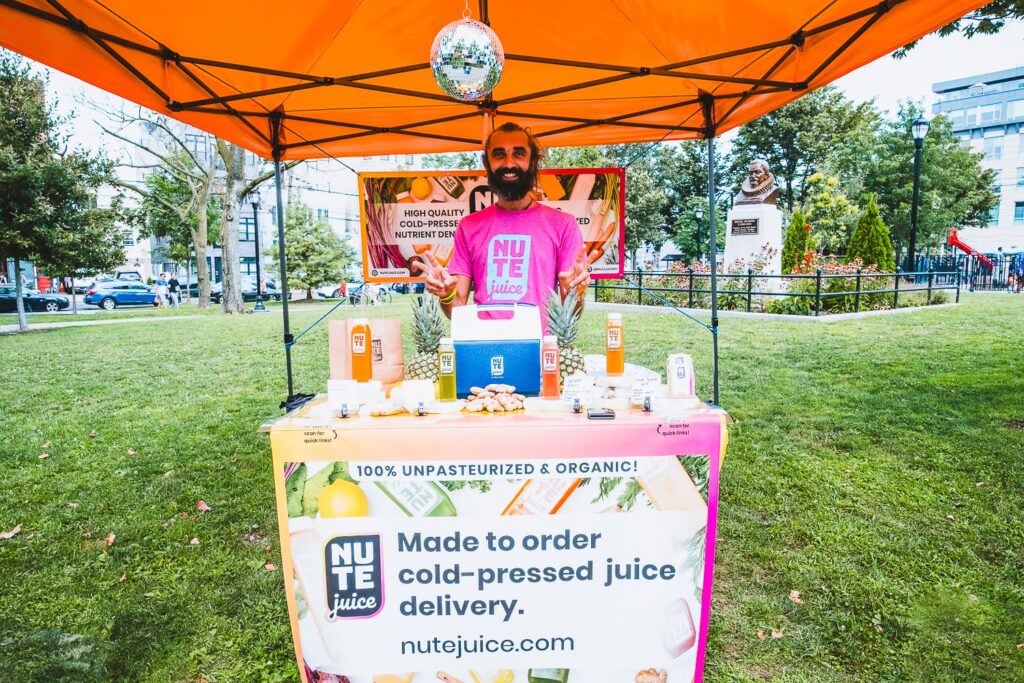 Photo of an White man, wearing a pink t-shirt, in a farmers market booth on a summer day, selling cold pressed juices