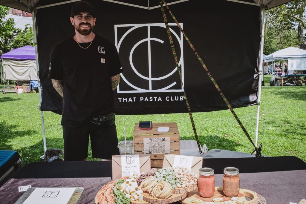 Photo of a smiling White man, wearing a black t-shirt & black jeans, in a farmers market booth on a summer day, selling fresh made pastas