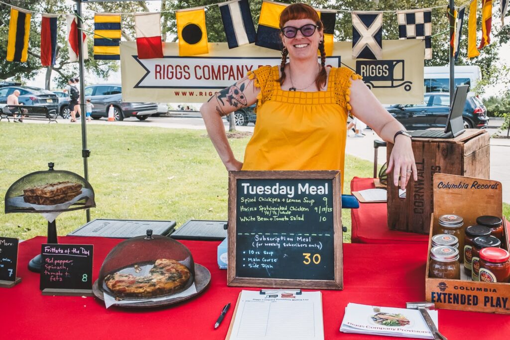 Photo of a smiling White woman, wearing a bright yellow shirt and glasses, in a farmers market booth on a summer day, selling prepared foods