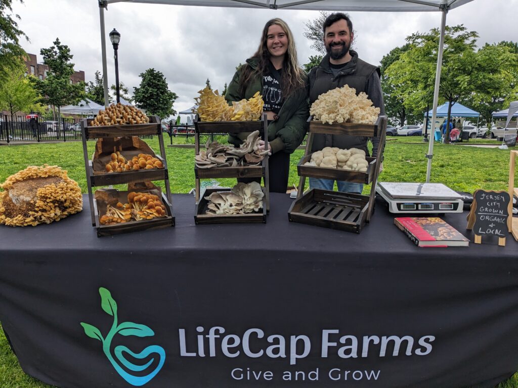 Photo of a couple, one man, one woman, at a farmers market booth selling various forms of mushrooms