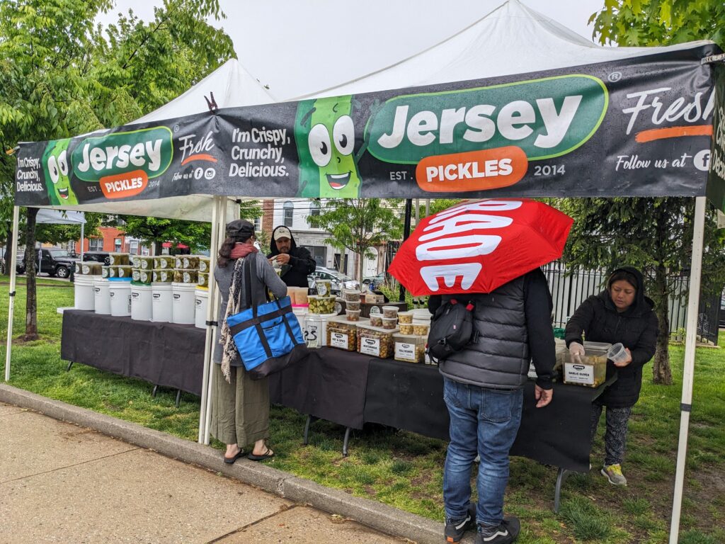 Photo of two people with their backs to the camera shopping at a farmers market booth selling pickles