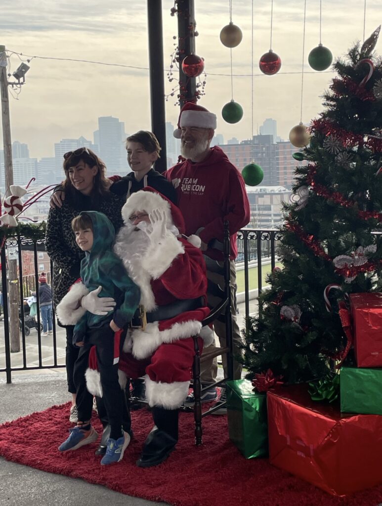 A family of four poses with Santa in an open-air gazebo next to a tree. Christmas ornaments hang in the background and the faint shadow of the NYC skyline can be seen in the distance.