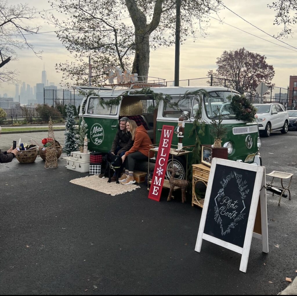 A green vintage VW bus is parked with the side door open and a couple sitting on the edge with their dog for a photo moment. The outside is decorated with Holiday accompaniments and the city skyline can be seen in the background.