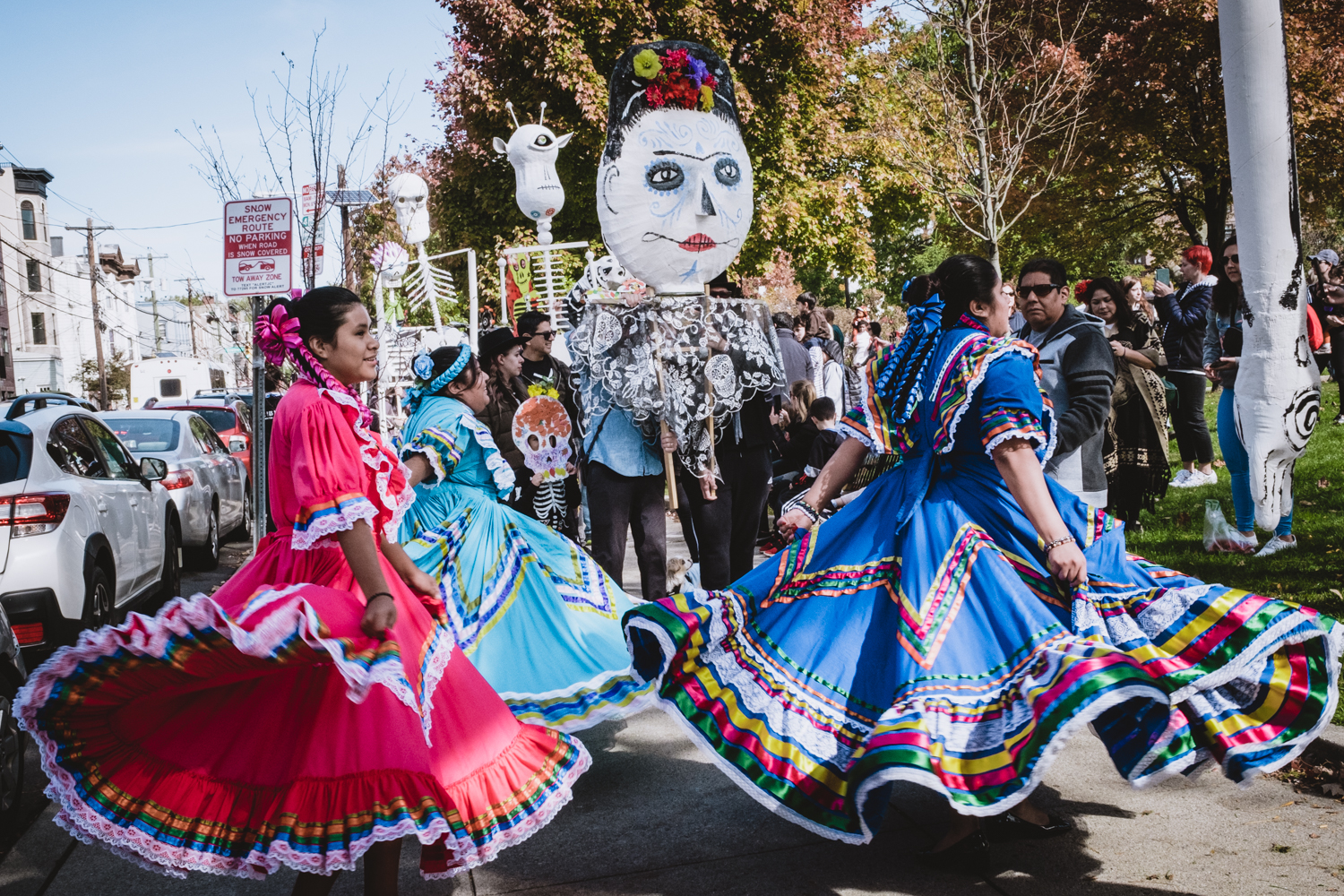 Three Mexican dancers dressed in traditional colorful attire dance and twirl their ornate skirts in front of the giant puppet parade procession.