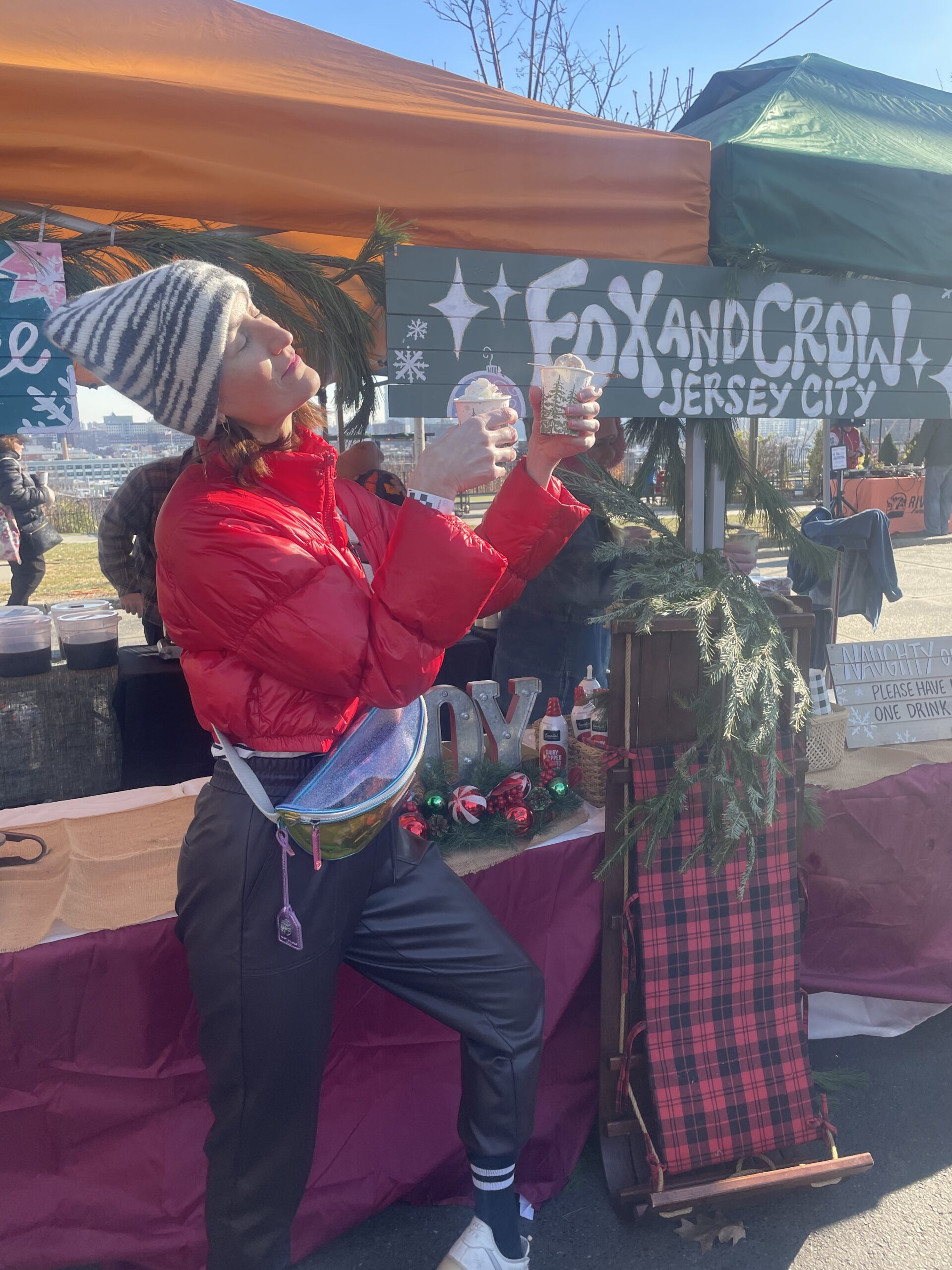 A woman in a red winter jacket and winter hat stands in front of the Fox & Crow stand at the Heights Holiday Market. Her face is turned towards the sun and she's holding two festive drinks in her hands.