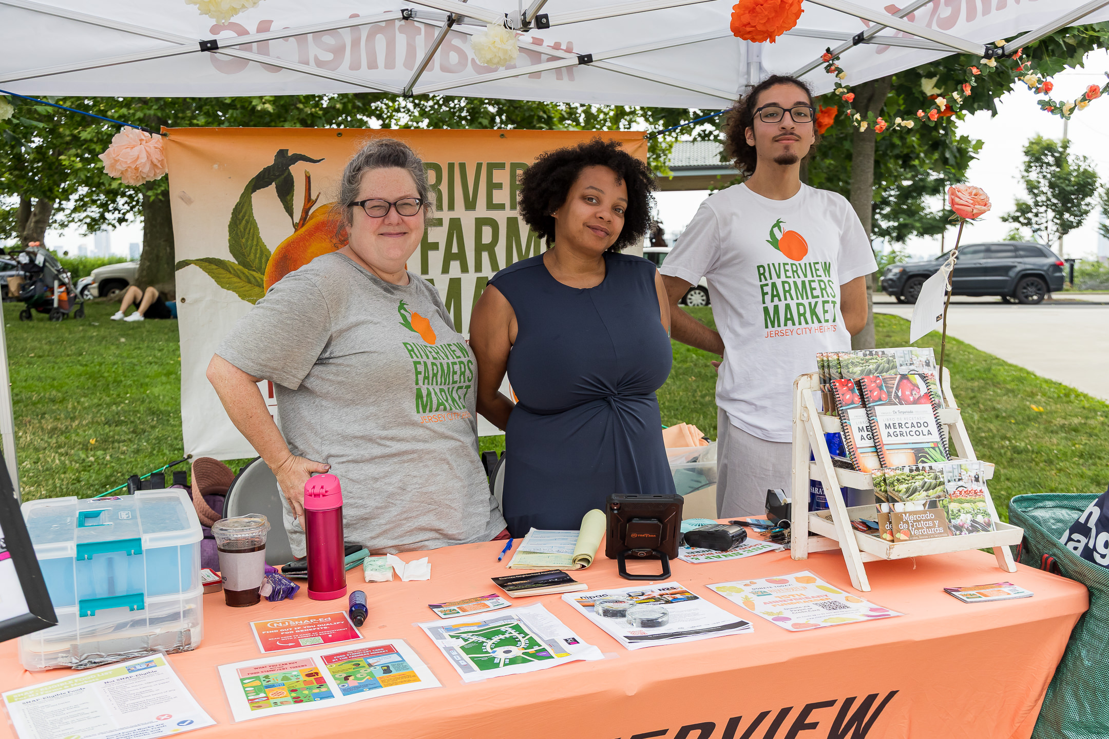 Our farmers market staff standing in a tent behind a table, the table has a peach-colored tablecloth on it, and is strewn with flyers and other materials.
