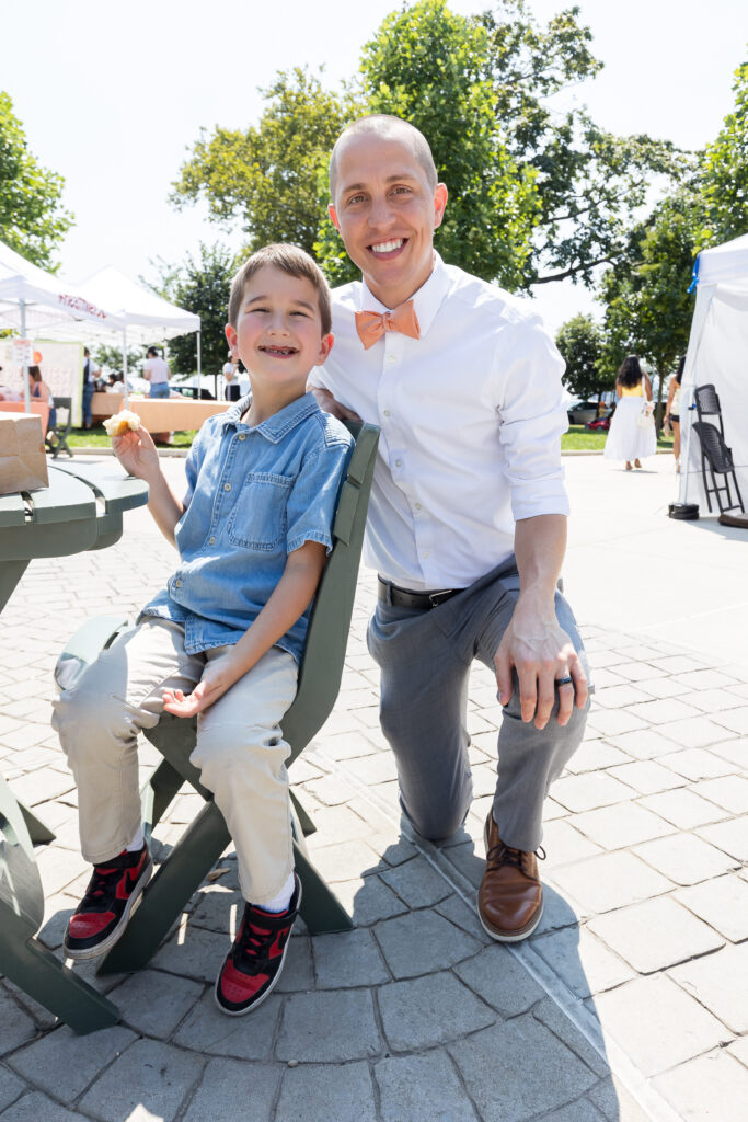 A man with a peach-colored bowtie kneels next to a seated child outdoors in the park.