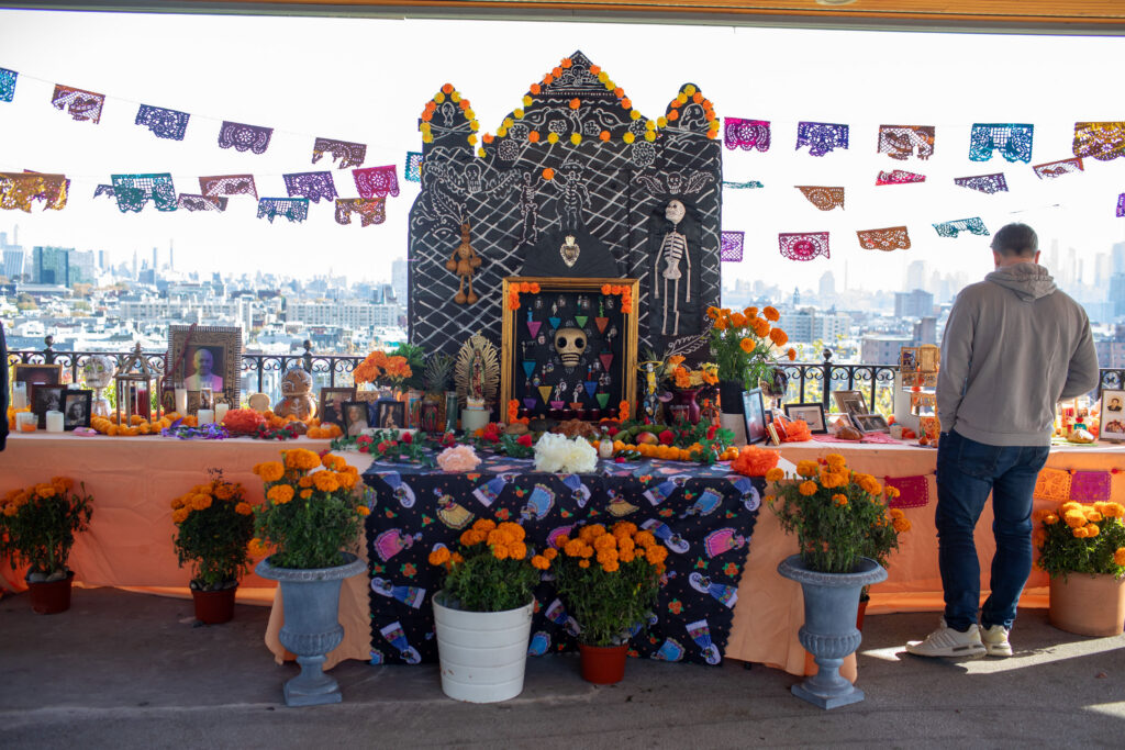The ofrenda or altar that was set up for the 2025 Day of the Dead Festival in Jersey City. A man stands in front of the altar with his back to the camera. The altar is decorated with frames and skeletons and many marigolds.